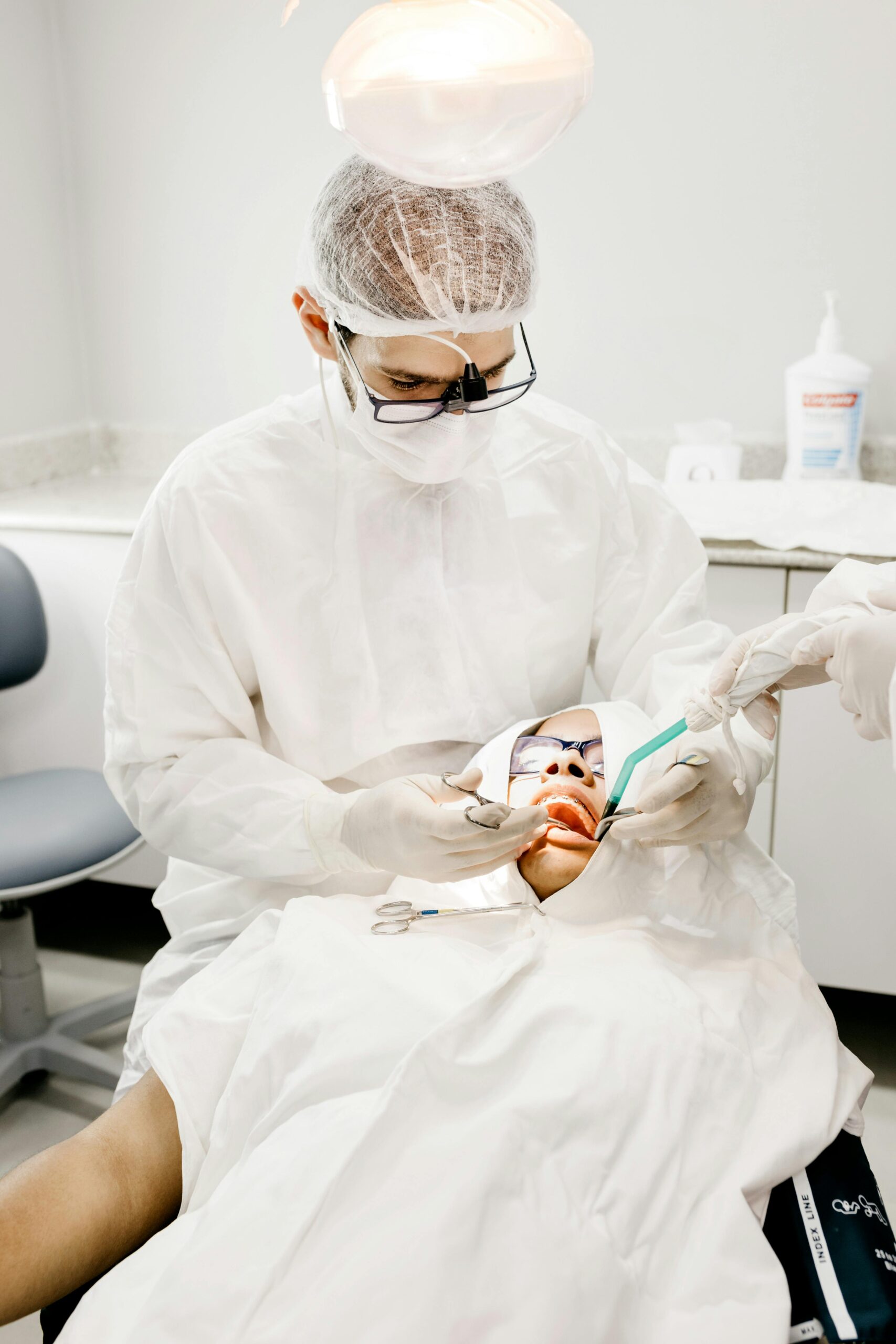 From above of unrecognizable orthodontist in mask and eyeglasses treating teeth of man with crop assistant using medical tools